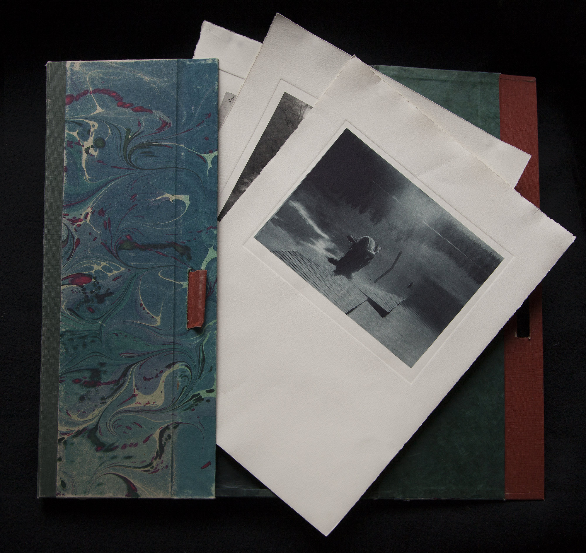 Open handmade portfolio displaying loose-edged photographic prints beside a marbled cover.