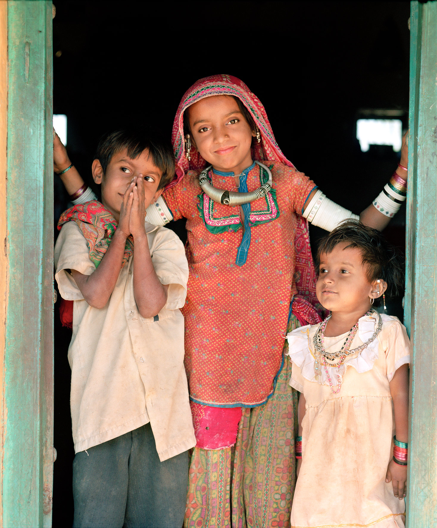 Three children standing in a doorway wearing traditional clothing and jewellery in Kutch, Gujarat.
