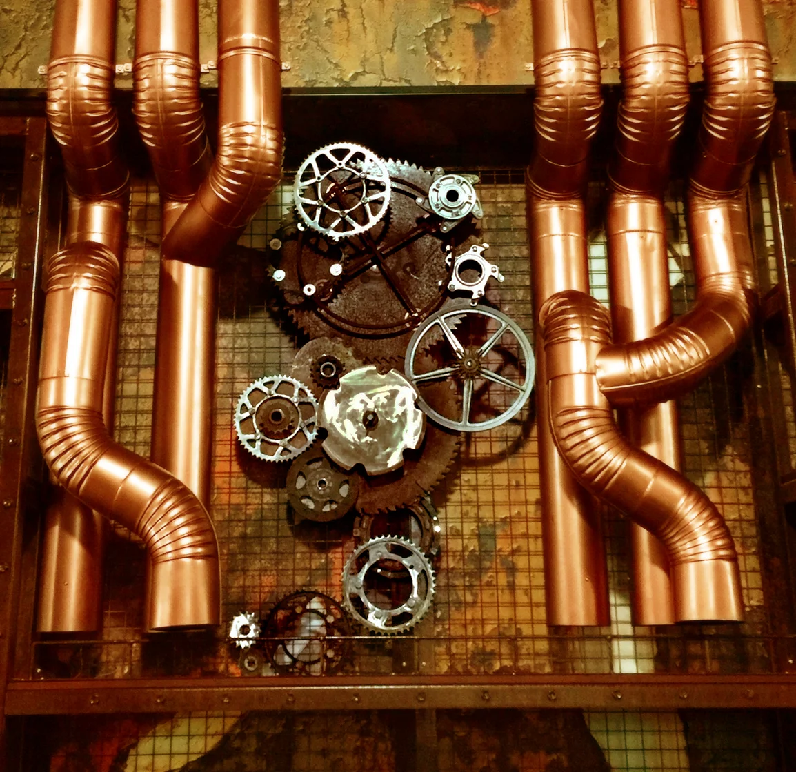 Detail of Machines from Hell by Magi Viljanen, showing gears and mechanical parts framed by large copper pipes.