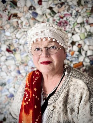 Portrait of Magi Viljanen in front of a wall covered with fragments of patterned ceramics.