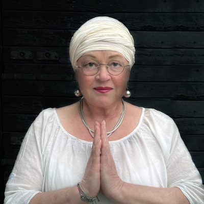 Magi Viljanen, self-portrait. The photographer is shown frontally in a white headwrap and blouse, hands together in a gesture of greeting or contemplation, against a dark wooden background.
