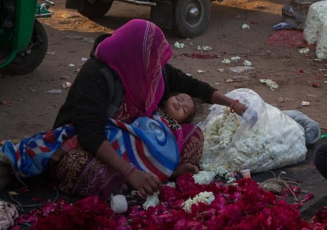 A flower seller sits among roses and jasmine blossoms while a child sleeps in her lap at Ahmedabad flower market.