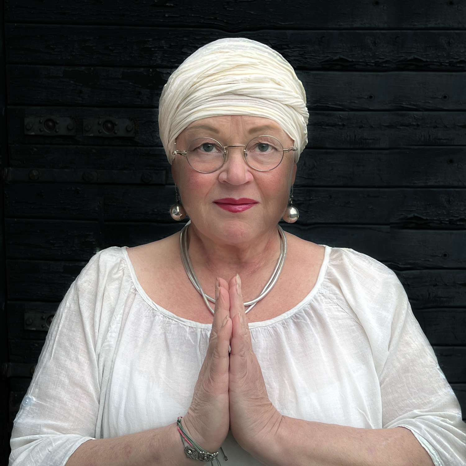 Magi Viljanen, self-portrait. The photographer is shown frontally in a white headwrap and blouse, hands together in a gesture of greeting or contemplation, against a dark wooden background.