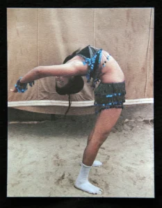 A young circus performer bends backwards in a contortion pose in front of a tent.