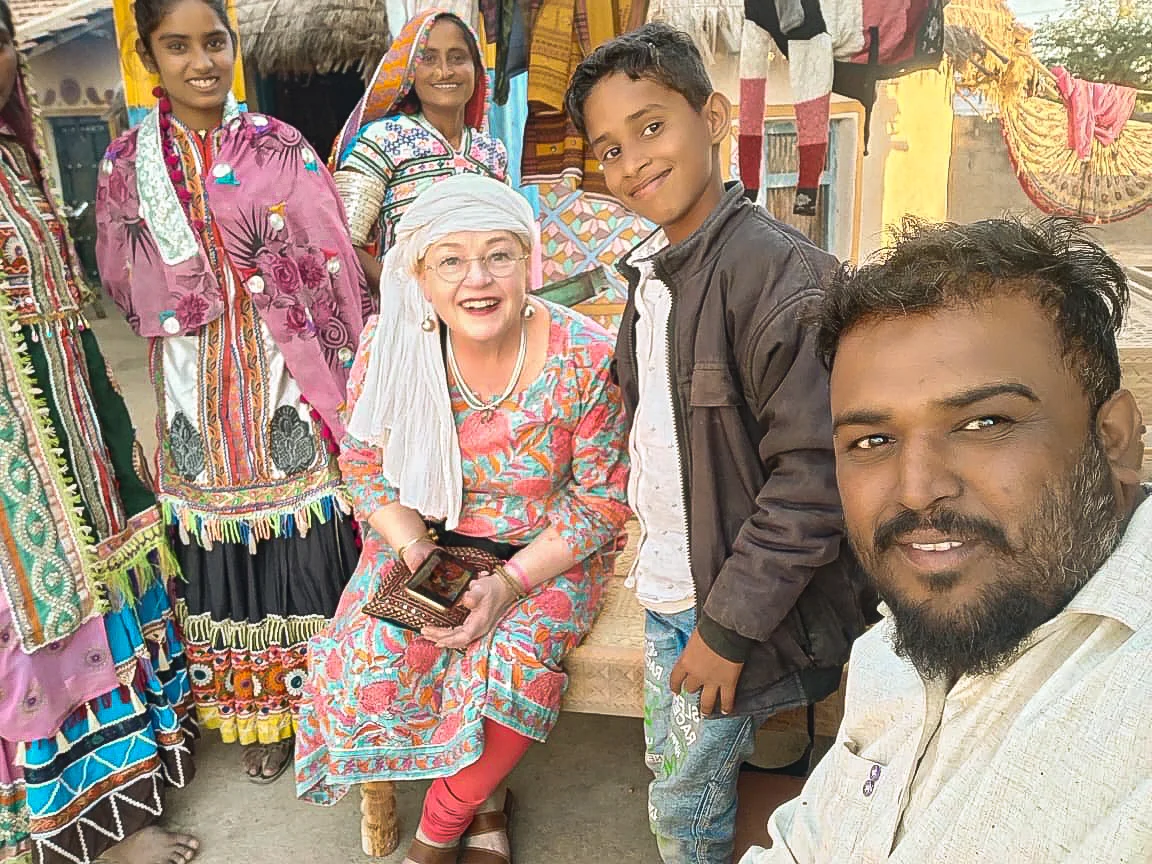 Magi Viljanen seated with a group of smiling people in India, several dressed in colourful, richly patterned traditional clothing.