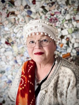 Portrait of Magi Viljanen in front of a wall covered with fragments of patterned ceramics.